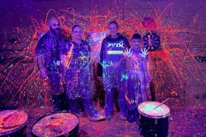 Four people in protective gear with neon paint splatters, standing in front of a dartboard under blacklight.
