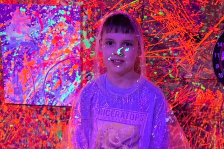 Person in clear poncho with neon paint splatters on wall and face glowing under blacklight.